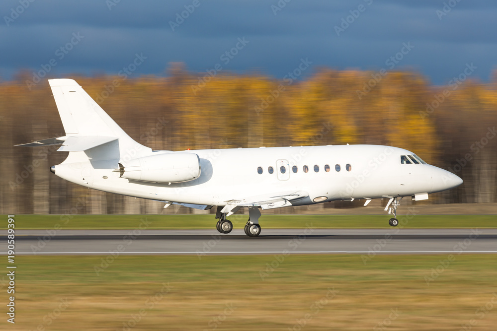 Side view of a luxury corporate or private jet landing in motion/ Landing business jet low over the runway, day light, blurred autumn forest on background/ Side view of a luxury corporate or private jet landing in motion/ Landing business jet low over the runway, day light, blurred autumn forest on background/