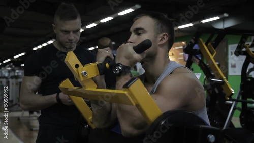 Male caucasian athlete is training his arms in the gym on the special equipment and his trainer is standing near. The sportsman is sitting on the special machine, holding the handles and benting his photo