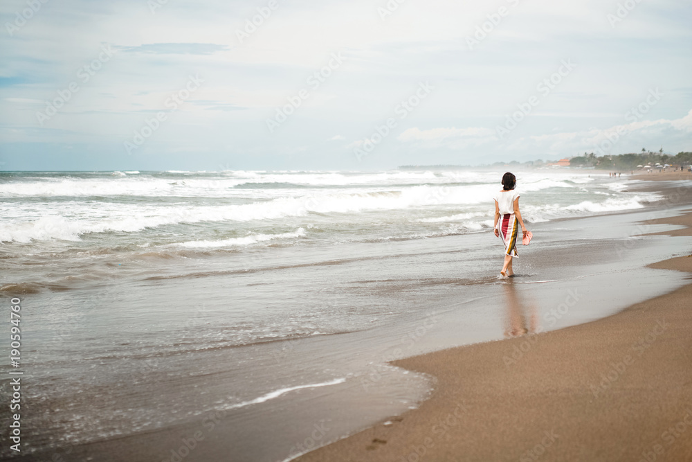 girl in a striped dress is walking along the beach of the ocean with gray sand and waves.