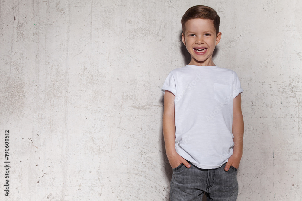 Cute little boy in white T-shirt posing in front of grunge concrete wall. Portrait of fashionable male child. Smiling boy posing, gray wall on background. Concept of children style and fashion. Cute little boy in white T-shirt posing in front of grunge concrete wall. Portrait of fashionable male child. Smiling boy posing, gray wall on background. Concept of children style and fashion.