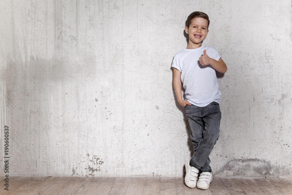 Cute little boy showing gesture thumb up. Portrait of smiling male child in gray trousers and white T-shirt. Positive little guy, gray wall on background, copy space. Cute little boy showing gesture thumb up. Portrait of smiling male child in gray trousers and white T-shirt. Positive little guy, gray wall on background, copy space.