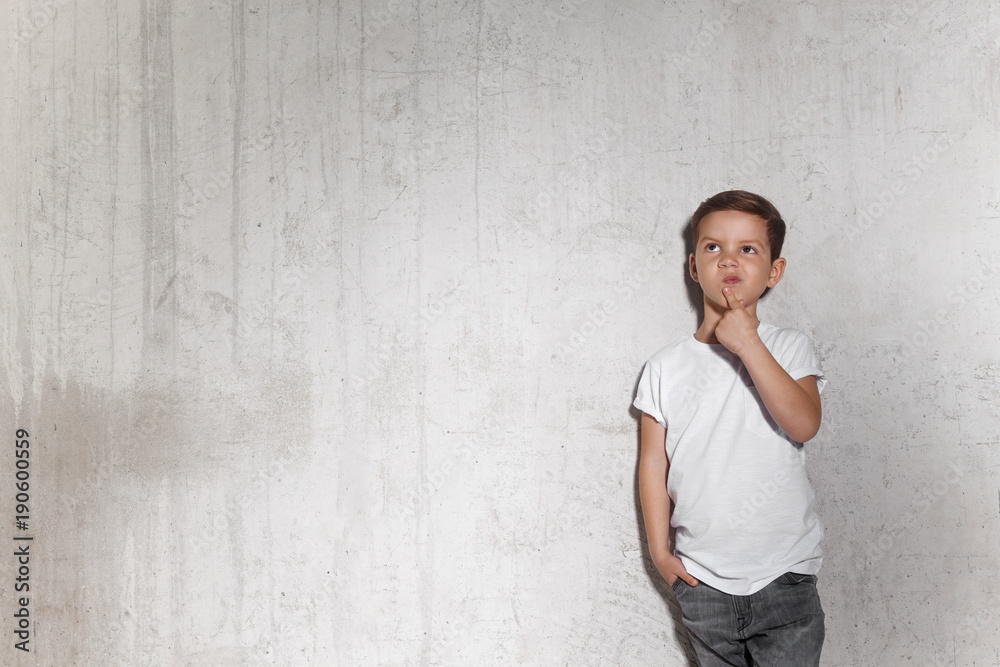 Pensive little guy stands on the wall background. Portrait of wistful boy in white T-shirt, copy space. Concept of reflections and looking for new idea. Pensive little guy stands on the wall background. Portrait of wistful boy in white T-shirt, copy space. Concept of reflections and looking for new idea.
