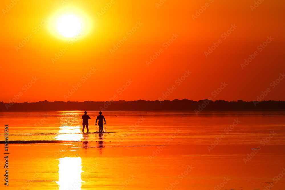 Silhouettes of people in shallow water in the lake with healing mud at orange red sunset