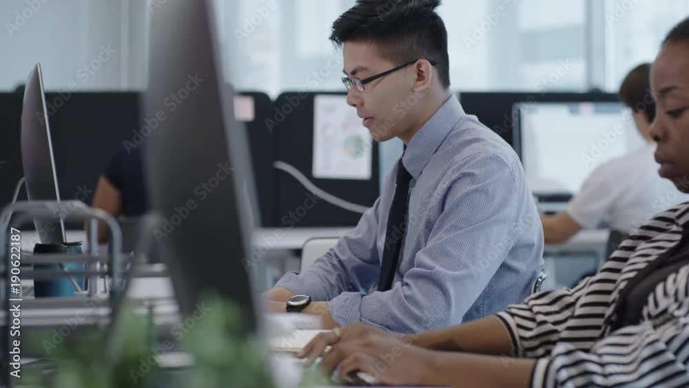 Busy office workers typing on computers in open space office; focus on young Asian clerk working at desk