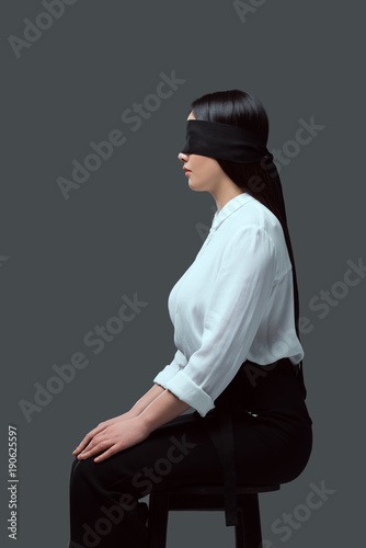 side view of young blindfolded woman sitting on chair isolated on grey