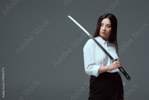 attractive young asian woman holding katana sword and looking away isolated on grey