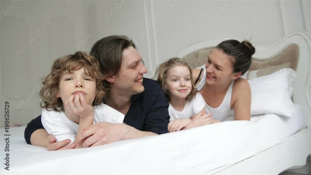 Portrait of Young Couple Spending Weekend with Their Children at Home. Little Boy and Girl with Two Adults are Relaxing Together on White Parents Bed. Portrait of Young Couple Spending Weekend with Their Children at Home. Little Boy and Girl with Two Adults are Relaxing Together on White Parents Bed.