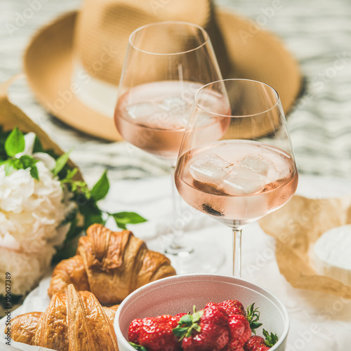French style romantic summer picnic setting. Flat-lay of glasses of rose wine with ice, fresh strawberries, croissants, brie cheese, straw hat, peony flowers, square crop. Outdoor gathering concept