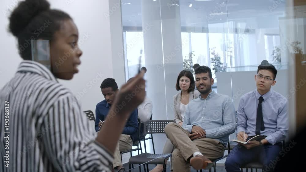 Diverse group of company employees sitting in glass meeting room and listening to African businesswoman at corporate training