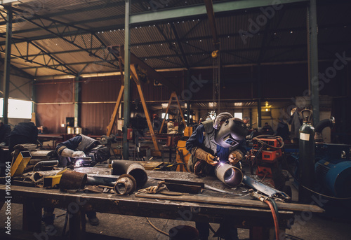Welder in protective uniform and mask welding metal pipe on the industrial table while sparks flying.