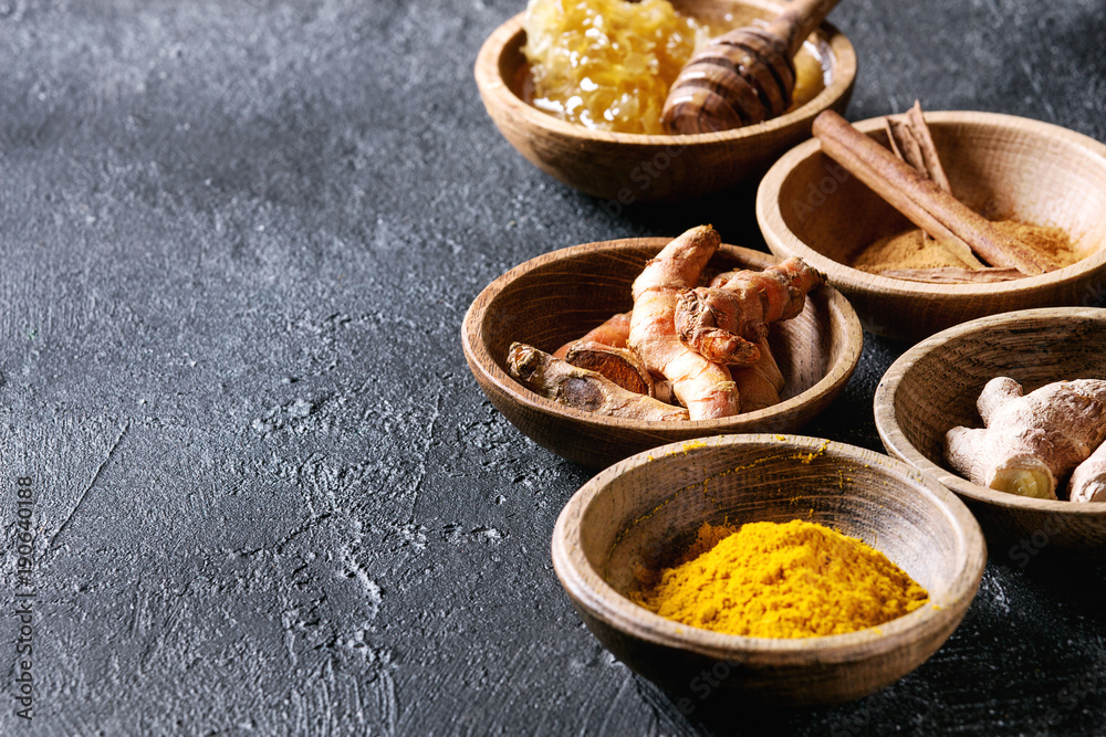 Fototapeta premium Ingredients for turmeric latte. Ground turmeric, curcuma root, cinnamon, ginger, honeycombs in wooden bowls over black texture background. Close up, copy space