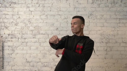 Young African guy in a black sweatshirt is dancing impetuously against a white wall. Disco, dance, movement, energy, youth concept. Lock at camera. 60 fps photo