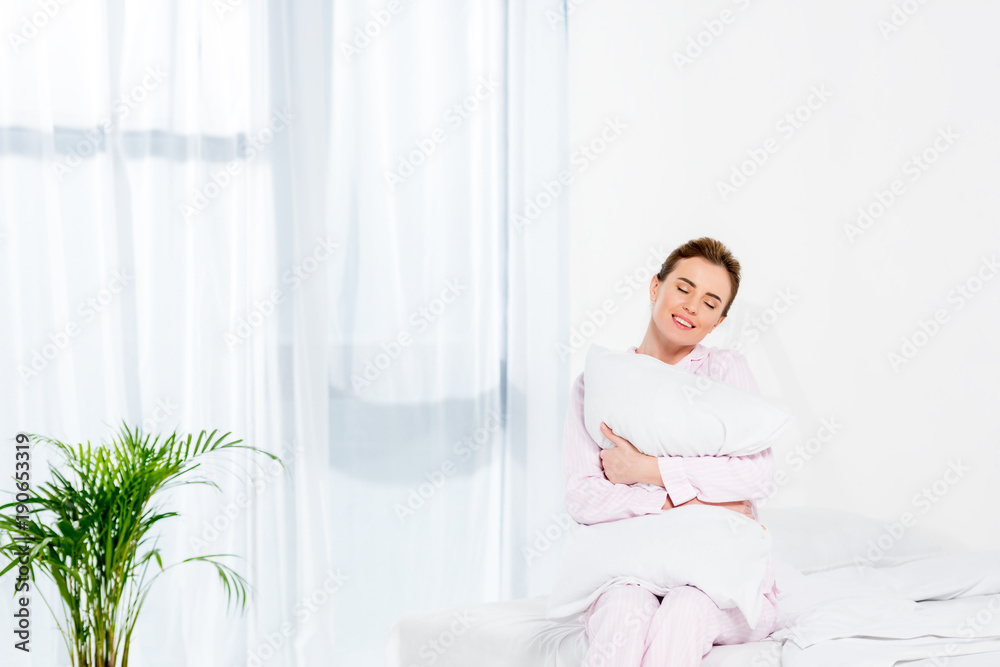 happy woman embracing pillow while sitting on bed in morning happy woman embracing pillow while sitting on bed in morning