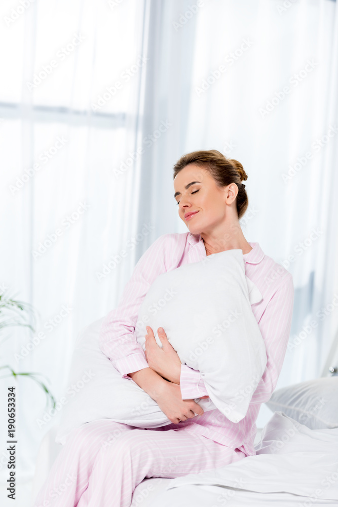 smiling woman embracing pillow while sitting on bed in morning smiling woman embracing pillow while sitting on bed in morning