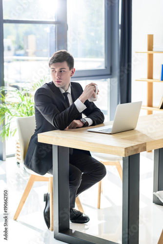 Young businessman drinks coffee while working on laptop in modern office