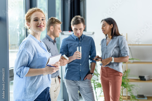 Young businesswoman with notepad in front of her colleagues discussing project in modern office