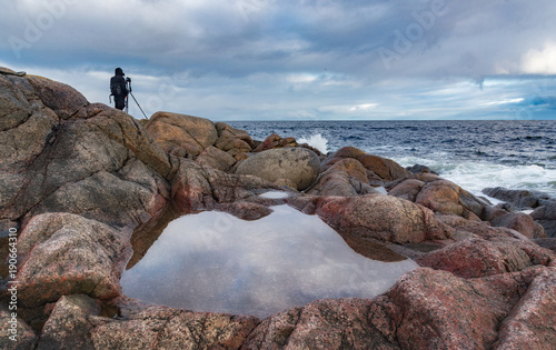 A lone figure of a photographer with a tripod on a stony ocean shore. With water among the stones in the foreground. Beautiful background for advertising photo tour. photo