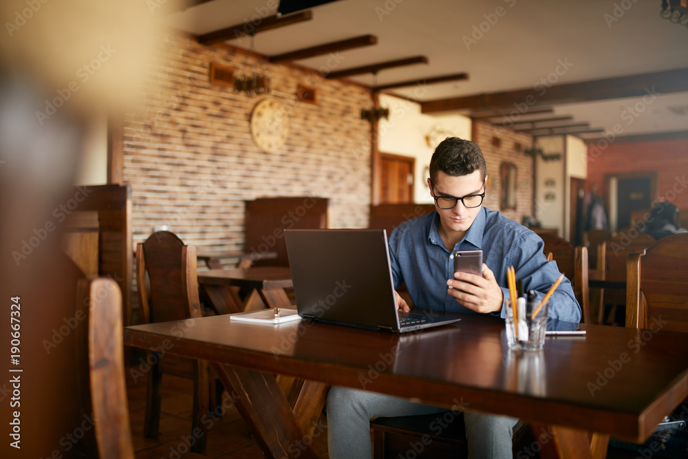 A young man typing a text on mobile modern smartphone. Hipster holding a modern phone and writing a phone message. Smiling young businessman in glasses looking at cellphone with laptop on table. A young man typing a text on mobile modern smartphone. Hipster holding a modern phone and writing a phone message. Smiling young businessman in glasses looking at cellphone with laptop on table.