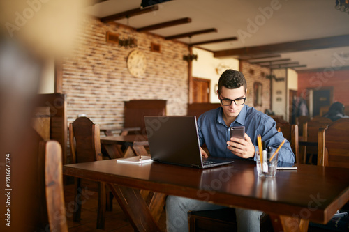 A young man typing a text on mobile modern smartphone. Hipster holding a modern phone and writing a phone message. Smiling young businessman in glasses looking at cellphone with laptop on table.