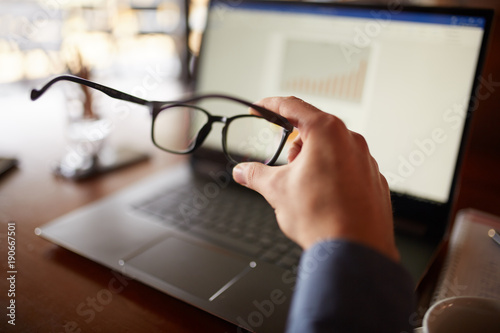 Close-up isolated shot of mans hand holding eyeglasses in front of laptop screen with charts and diagrams. Poor eyesight threatment theme. Computer glasses. photo