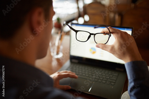 Back view of man hand holding eyeglasses in front of laptop screen with charts and diagrams. Poor eyesight threatment theme. Computer glasses. Break for eye rest. photo