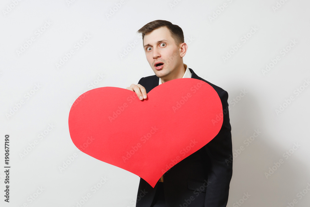 Young stunned shocked upset sad man in suit holding big red heart isolated on white background. Copy space for advertisement. St. Valentine's Day, International Women's Day, birthday, holiday concept. Young stunned shocked upset sad man in suit holding big red heart isolated on white background. Copy space for advertisement. St. Valentine's Day, International Women's Day, birthday, holiday concept.