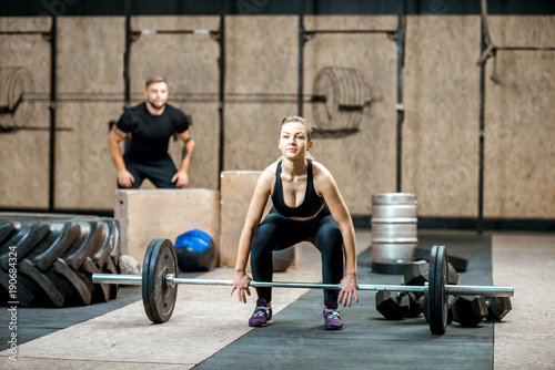 Young athletic woman lifting up a burbell with man training on the background in the crossfit gym