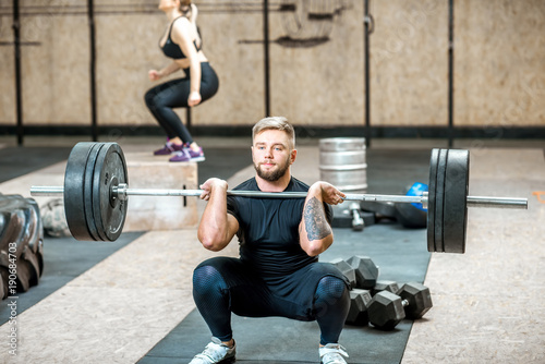 Handsome athletic man in black sports wear lifting up a heavy burbell with woman training on the background in the crossfit gym