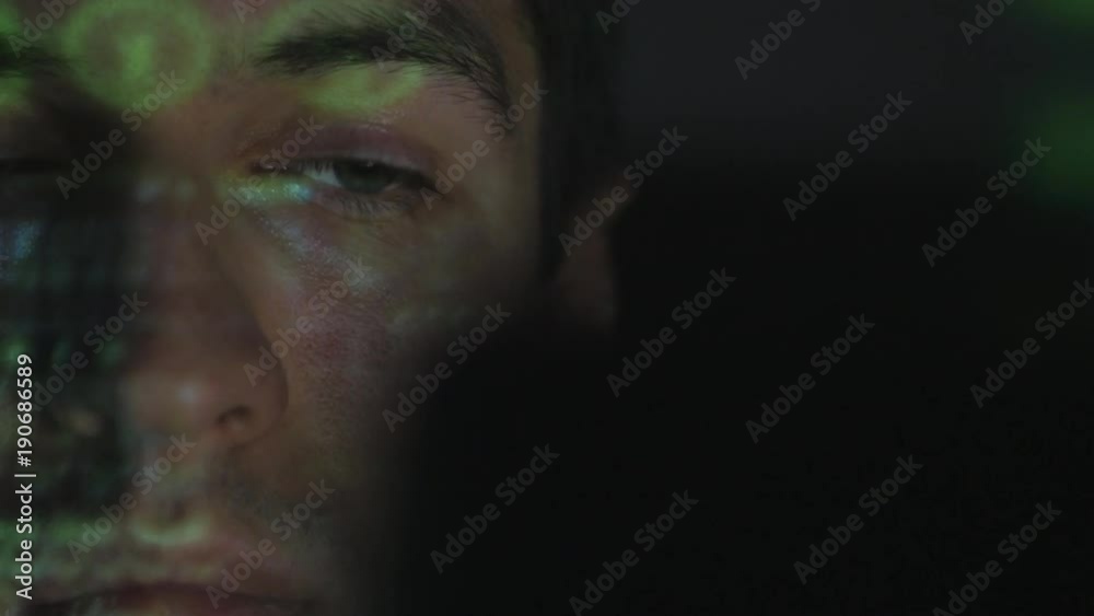 Close up Man programmer hacker working on a computer while green code characters reflect on his face in a dark office room.