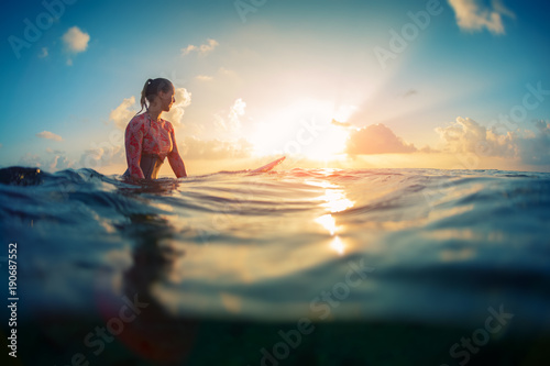 Young lady surfer waits the waves in ocean during sunrise with surf board.