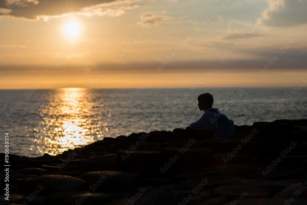 Sunset at sea side, light reflection on water surface, little boy sits at the beach, body and face silhuoette