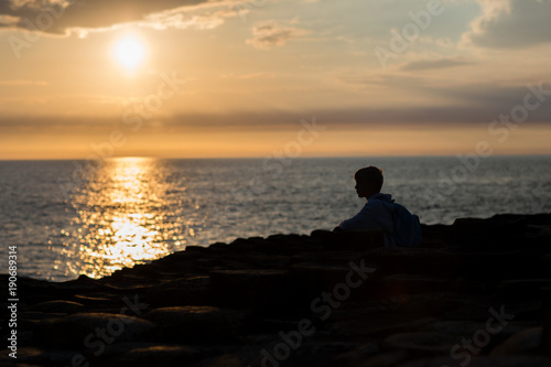 Sunset at sea side, light reflection on water surface, little boy sits at the beach, body and face silhuoette