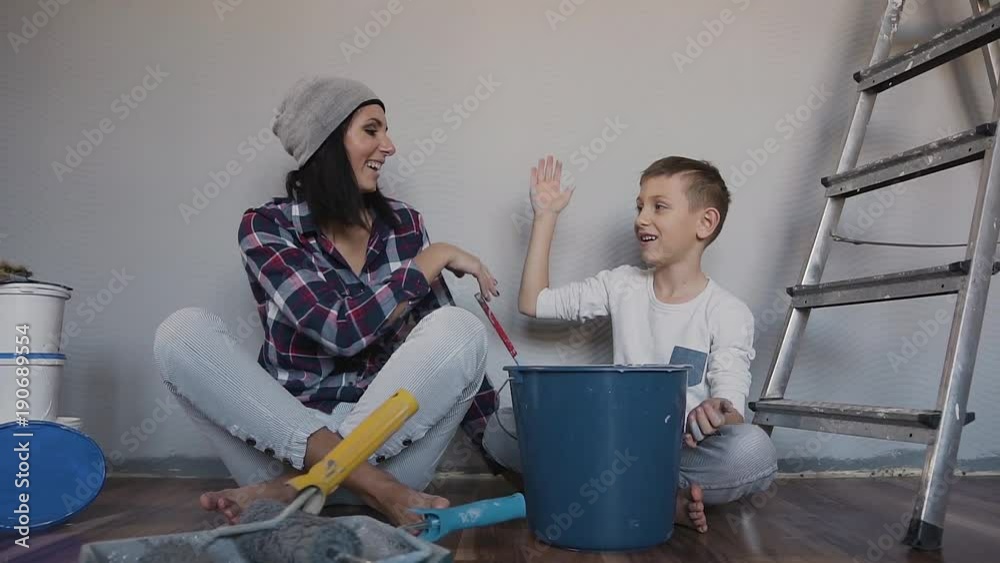 The boy and his mother are sitting on the floor, looking at the room that they just painted in gray color and joyfully giving five to each other The boy and his mother are sitting on the floor, looking at the room that they just painted in gray color and joyfully giving five to each other