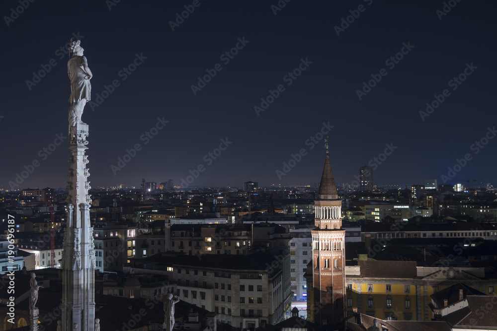 Night view of Milan (Italy) seen from the roof of the cathedral, a spire with a marble statue in the foreground and the city in the background. Italian cityscape.