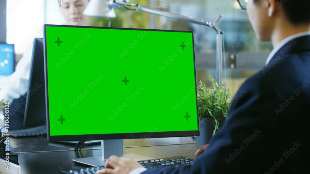 In the Office Businessman works on a Desktop Personal Computer with Mock-up Green Screen. Colleague Enters Office and Takes Place at Her Desk.