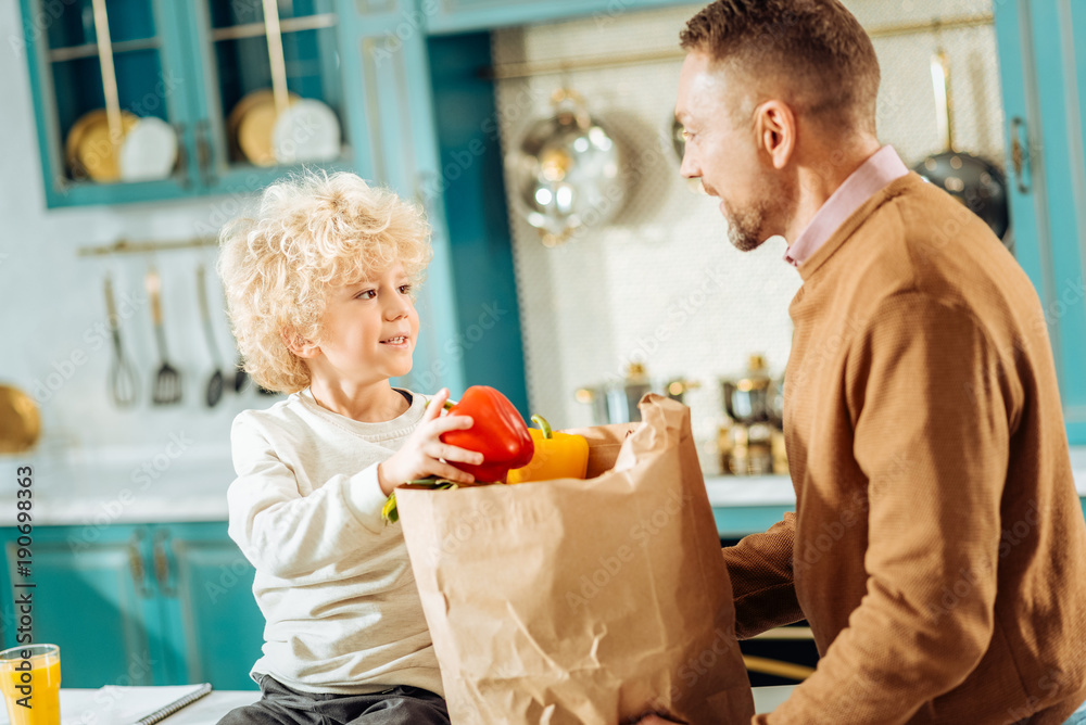Father and son. Cheerful cute nice boy holding pepper and looking at his father while standing with him in the kitchen Father and son. Cheerful cute nice boy holding pepper and looking at his father while standing with him in the kitchen