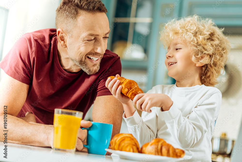 Wonderful mood. Happy delighted cute boy holding a croissant and laughing while talking to his father Wonderful mood. Happy delighted cute boy holding a croissant and laughing while talking to his father