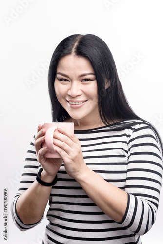 Delicious coffee. Cute dark-haired woman cupping a mug of coffee in her hands and smiling at the camera while posing isolated on a white background