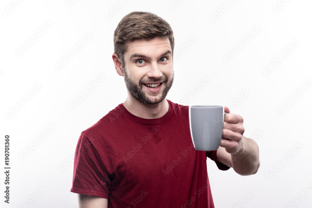 Give it a try. Pleasant young man in a t-shirt offering a cup of coffee and smiling at the camera while posing isolated on a white background