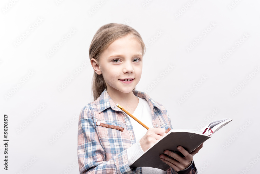 Preparation for school. Cute little girl writing in a notebook with a pencil and smiling at the camera while posing isolated on a white background Preparation for school. Cute little girl writing in a notebook with a pencil and smiling at the camera while posing isolated on a white background