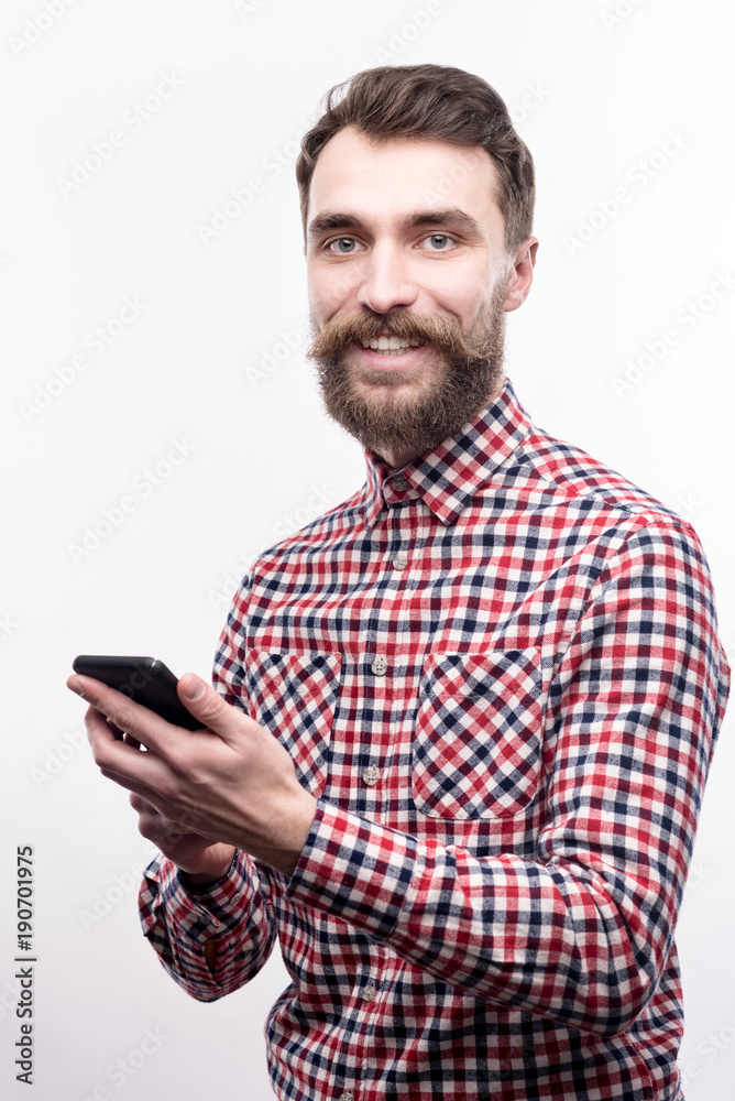 Pleasant communication. Cheerful bearded young man in a checked shirt texting someone via his phone and smiling happily at the camera while posing isolated on a white Pleasant communication. Cheerful bearded young man in a checked shirt texting someone via his phone and smiling happily at the camera while posing isolated on a white