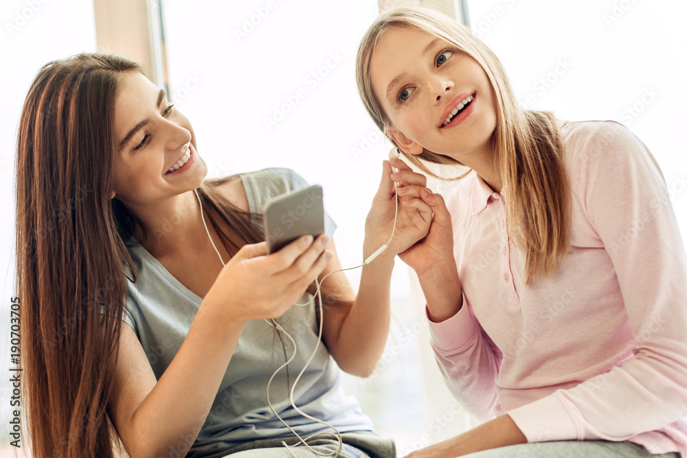 Sharing inspiration. Charming teenage girl sitting on the windowsill together with her younger sister and giving one earphone to her while listening to music Sharing inspiration. Charming teenage girl sitting on the windowsill together with her younger sister and giving one earphone to her while listening to music