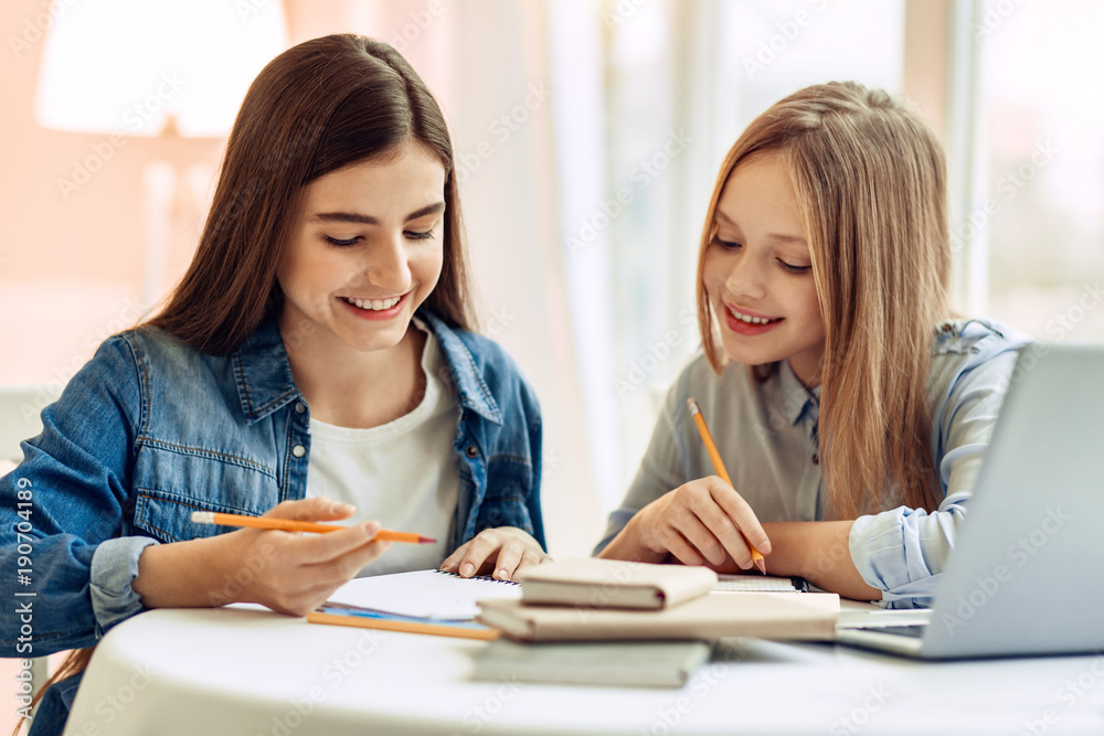 Pleasant cooperation. Upbeat teenage girls sitting at the table and doing their home assignment, helping each other with it while smiling Pleasant cooperation. Upbeat teenage girls sitting at the table and doing their home assignment, helping each other with it while smiling