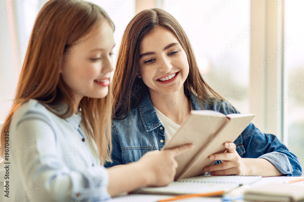 Knowledge is power. Charming teenage girls sitting at the table in the living room and reading a book together while doing their home assignment Knowledge is power. Charming teenage girls sitting at the table in the living room and reading a book together while doing their home assignment