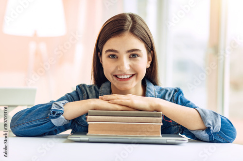 Love learning. Pretty upbeat teenage girl sitting at the table and resting her hands on a pile of study materials while posing for the camera © Viacheslav Yakobchuk