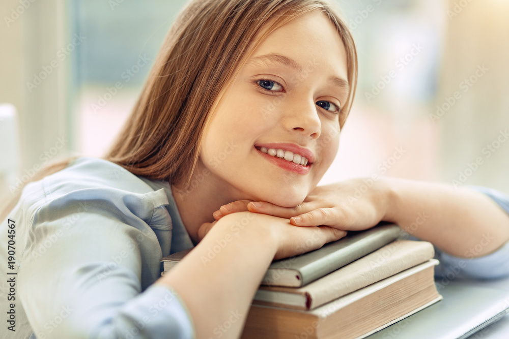 Relaxed student. Beautiful teenage girl posing for the camera while resting her chin on her hands folded on a pile of books and smiling Relaxed student. Beautiful teenage girl posing for the camera while resting her chin on her hands folded on a pile of books and smiling