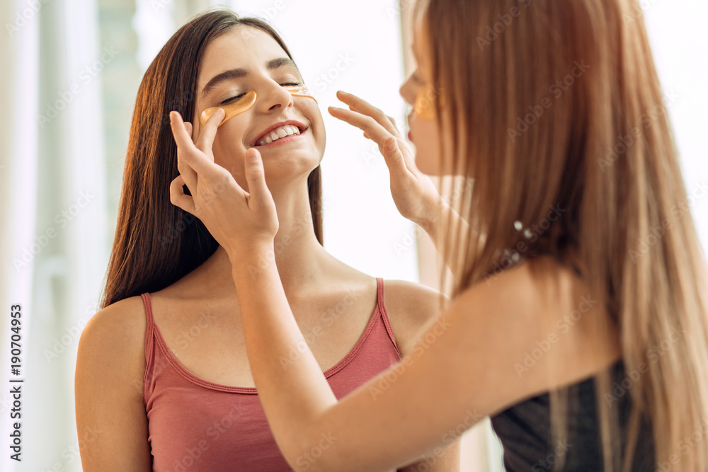 Sisterly care. Pleasant teenage girl helping her elder sister apply under-eye patches, adjusting them properly on her skin while the other girl smiles Sisterly care. Pleasant teenage girl helping her elder sister apply under-eye patches, adjusting them properly on her skin while the other girl smiles
