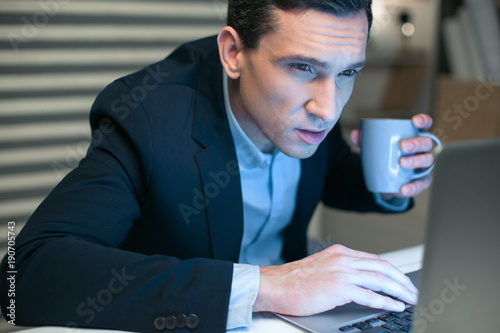 Computer error. Serious ambitious pretty businessman carrying cup while working on laptop and staring at the screen