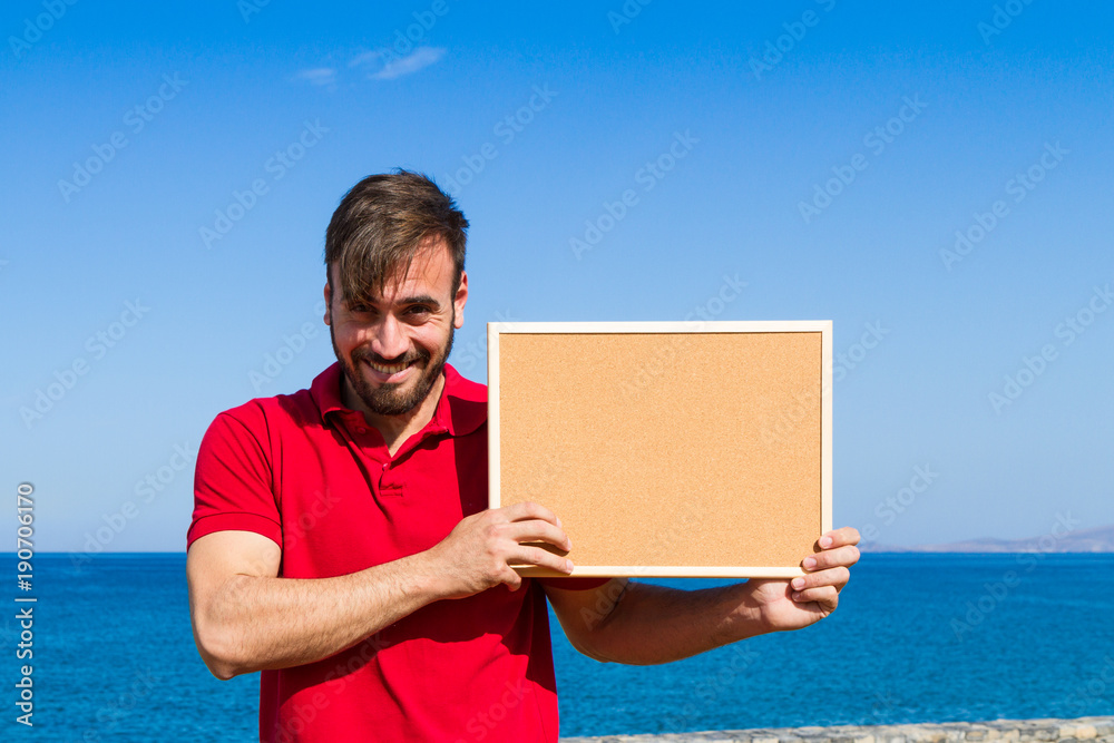 Bearded attractive inviting young man holding a framed blank corkboard in blue sky Bearded attractive inviting young man holding a framed blank corkboard in blue sky