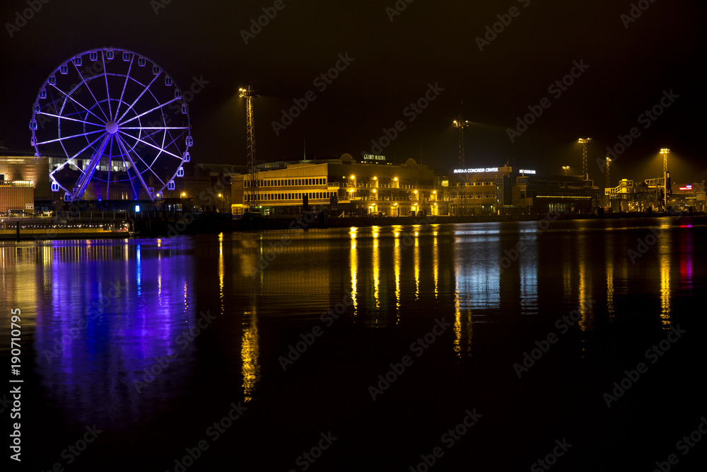 Panorama of the waterfront in Helsinki, Finland, during the long winter night. A ferris wheel and the yellow lights of the buildings are reflected on the water of the sea